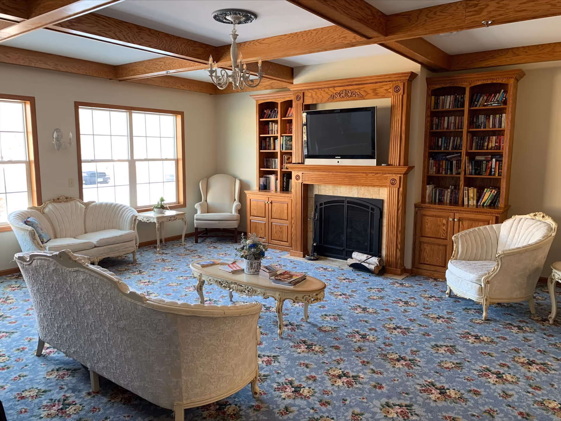 A cozy senior living room with floral carpet, antique-style cream upholstered sofas and chairs, a wooden coffee table with magazines and a flower arrangement, large windows letting in natural light, and a wooden built-in bookshelf and fireplace with a mounted flat-screen TV.