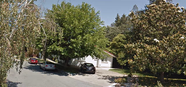 A quiet residential street with several parked cars and a white single-story building partially visible behind large green trees and bushes on a sunny day.