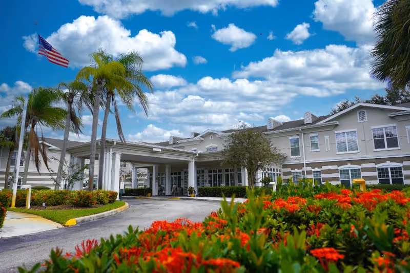 Exterior view of Sodalis Stuart facility with a driveway leading to the entrance, surrounded by palm trees and vibrant red flowers under a partly cloudy blue sky. An American flag is flying on a flagpole near the entrance.