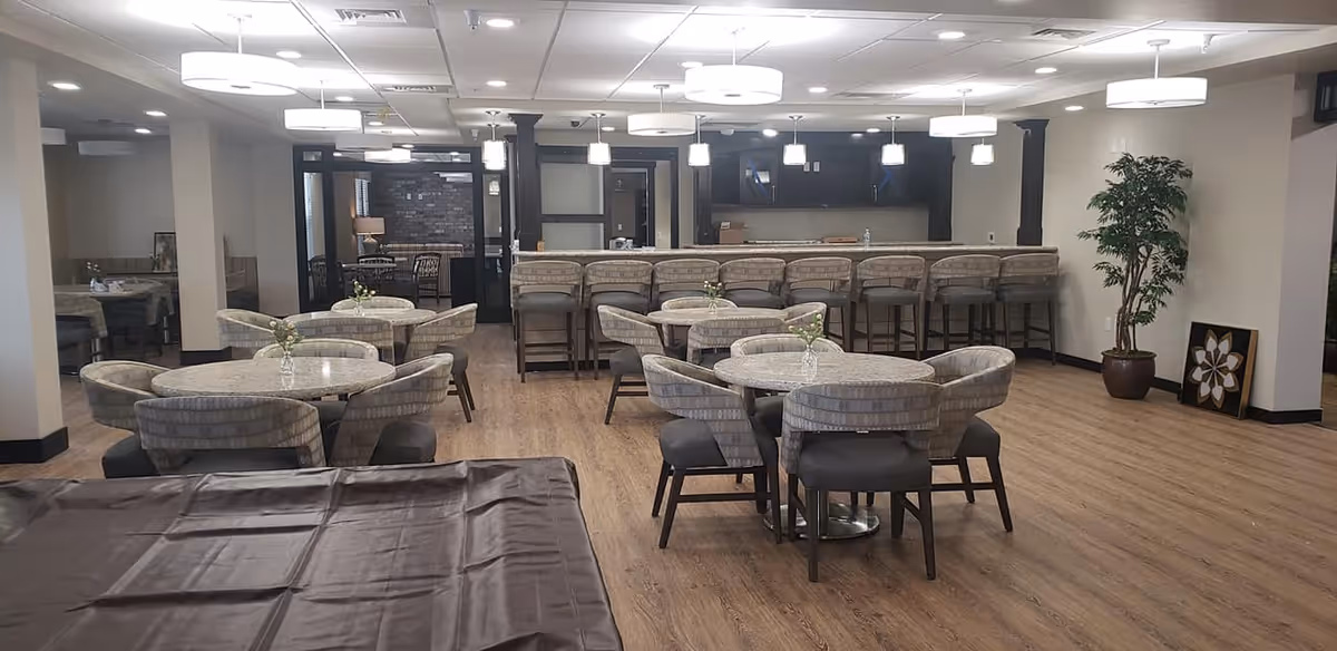 A spacious dining area with round tables and cushioned chairs arranged neatly. In the background, there is a long counter with high stools and pendant lights hanging above. The room has wooden flooring, modern ceiling lights, a potted plant in the corner, and a piece of framed artwork resting against the wall.