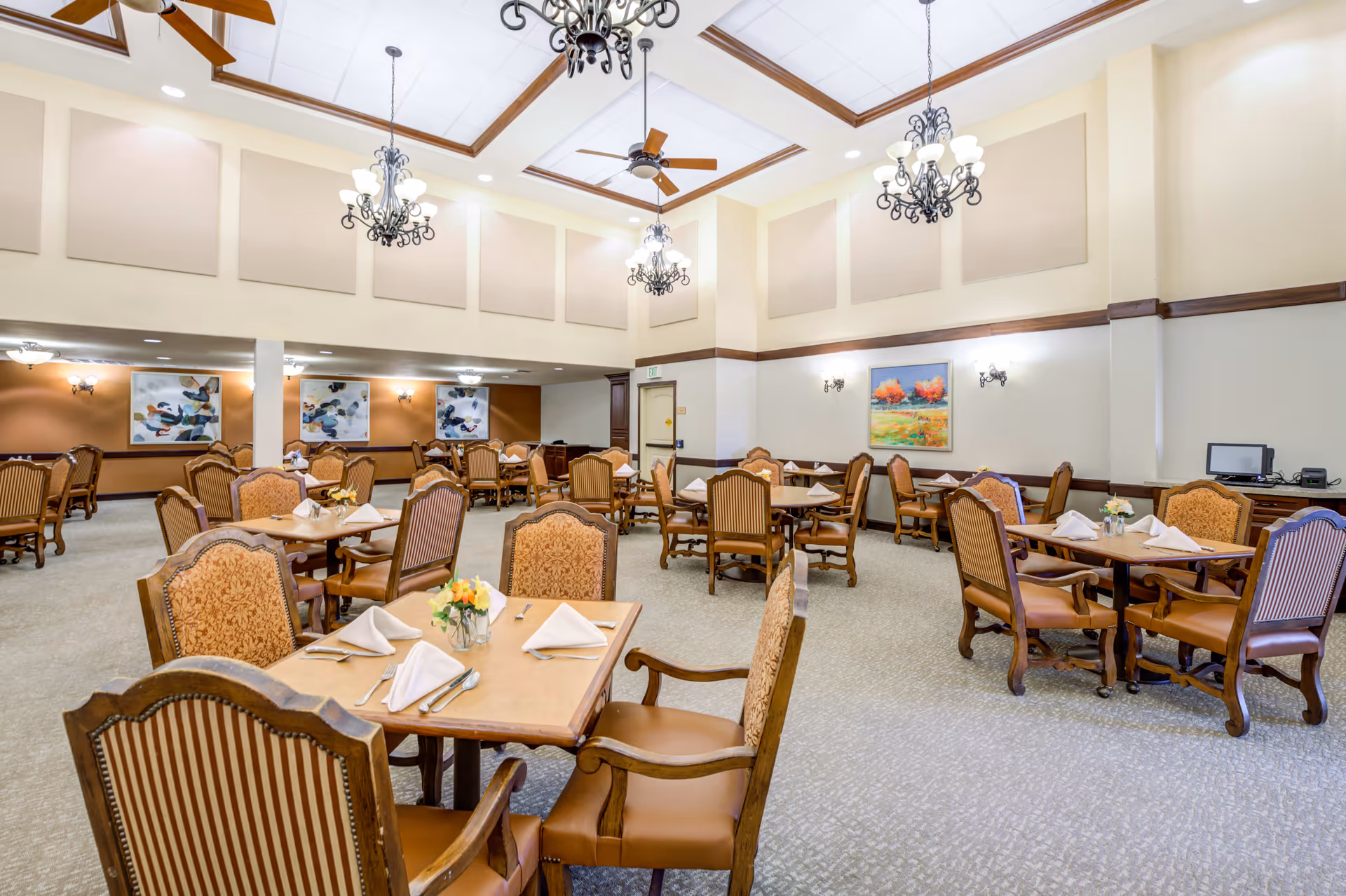 A spacious dining room with multiple wooden tables and upholstered chairs arranged neatly. Each table is set with folded white napkins, silverware, and small floral centerpieces. The room features high ceilings with decorative wooden beams, ceiling fans, and ornate chandeliers. The walls are adorned with framed paintings and soft lighting fixtures, creating a warm and inviting atmosphere.
