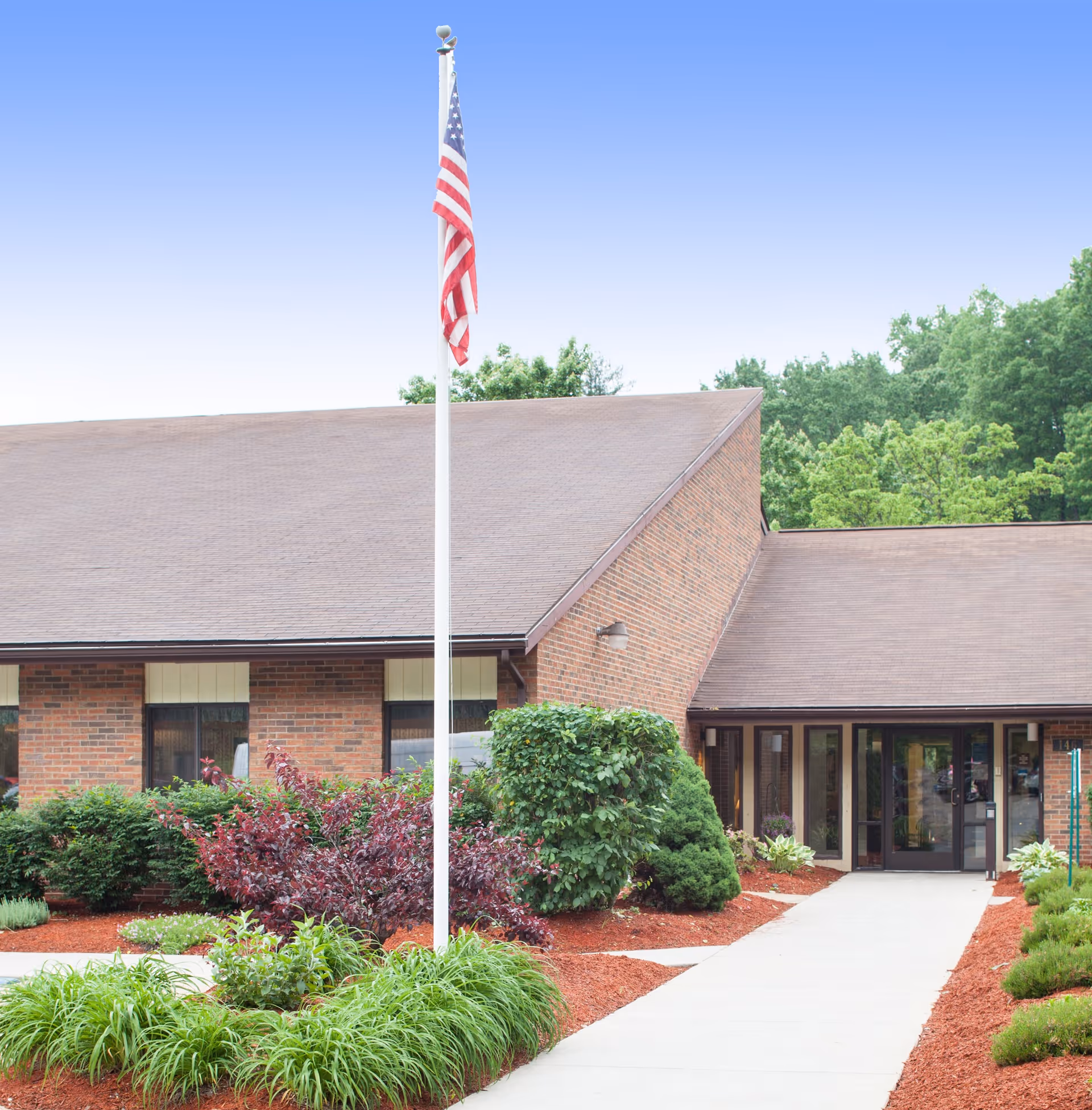 Brick building front with a flagpole displaying an American flag, landscaped beds and a paved walkway leading to glass entrance doors.