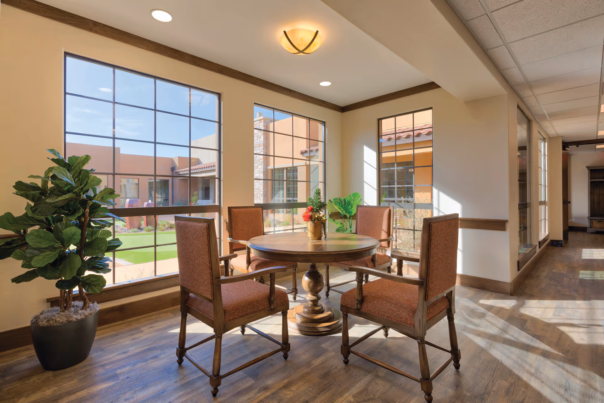 Sunlit seating area with a round wooden table and four upholstered chairs by large windows overlooking a courtyard.
