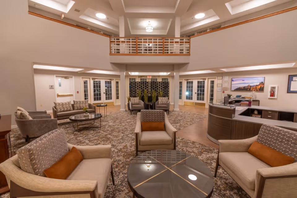 Spacious and well-lit senior living facility common area with comfortable armchairs and sofas arranged around glass coffee tables on a patterned carpet. The room features a high ceiling with recessed lighting, a balcony railing on the upper level, and a reception desk to the right. French doors and plants are visible in the background.