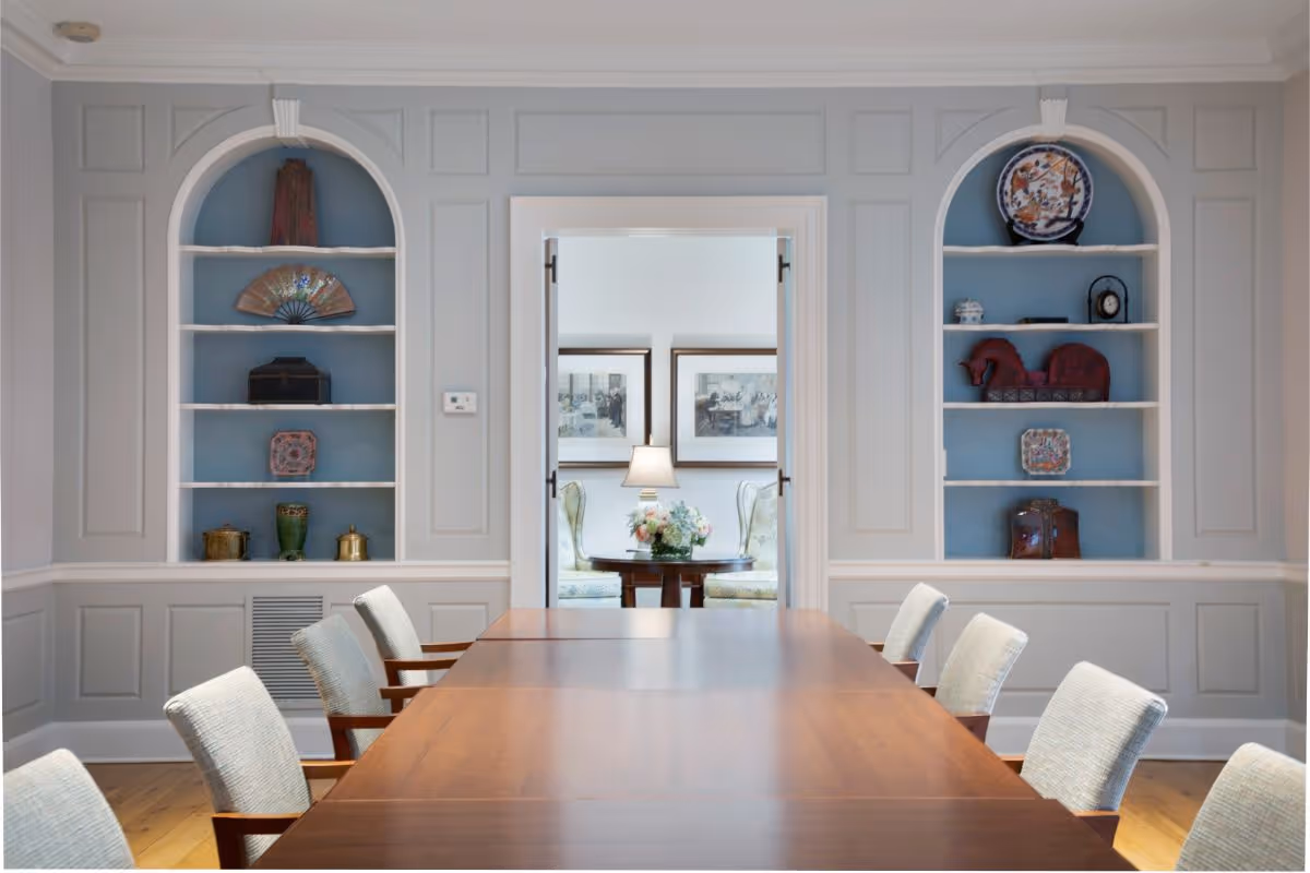 A formal dining room with a long wooden table surrounded by cushioned chairs. The walls are painted light gray with built-in shelves on either side of a doorway. The shelves contain decorative items including vases, plates, and small sculptures. Through the doorway, a small sitting area with two upholstered chairs, a round table, a lamp, and framed artwork on the wall is visible.
