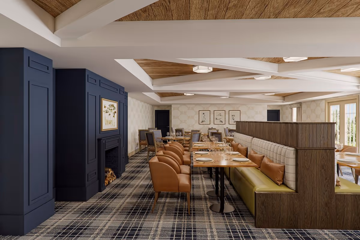 A dining room with a modern and cozy design featuring a long wooden table with place settings, cushioned chairs, and a built-in bench with pillows. The room has a patterned carpet, a dark blue fireplace with stacked firewood, light-colored walls with framed artwork, and a ceiling with white beams and wood paneling.