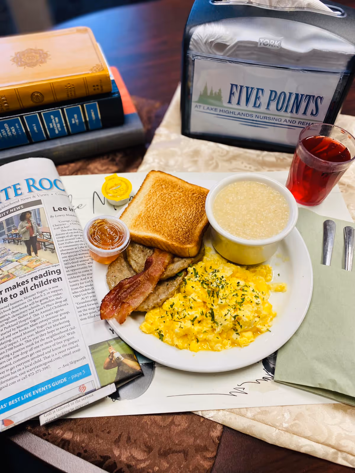 A breakfast plate with scrambled eggs garnished with herbs, two sausage patties, a strip of bacon, a slice of toasted bread, and a bowl of grits. Next to the plate is a small container of jelly, a yellow packet of butter, a glass of red juice, a folded green napkin with a fork and knife, and a newspaper. In the background, there is a napkin holder with a sign that reads 'Five Points at Lake Highlands Nursing and Rehab.'
