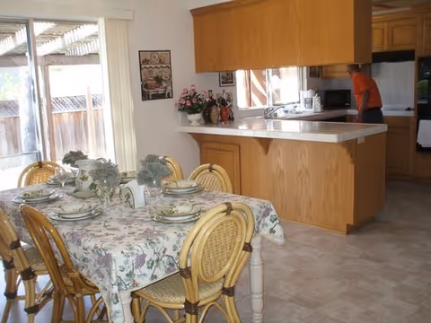 A dining area with a table covered by a floral tablecloth set with plates, bowls, and glasses. Four wicker chairs surround the table. In the background, there is a kitchen with wooden cabinets, a countertop, and a person standing near the sink. A sliding glass door with vertical blinds leads to an outdoor area.