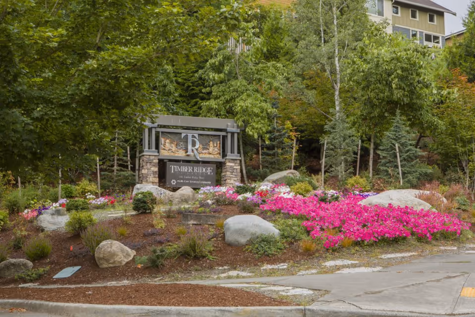 Landscaped entrance sign reading "Timber Ridge" surrounded by flowering beds, rocks, and trees with part of a building visible behind.