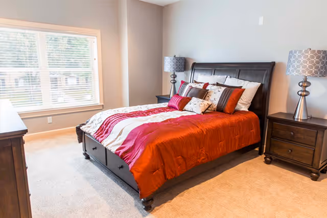 Sunlit bedroom with a wooden bed dressed in red and white bedding, matching nightstands and lamps, and a large window.