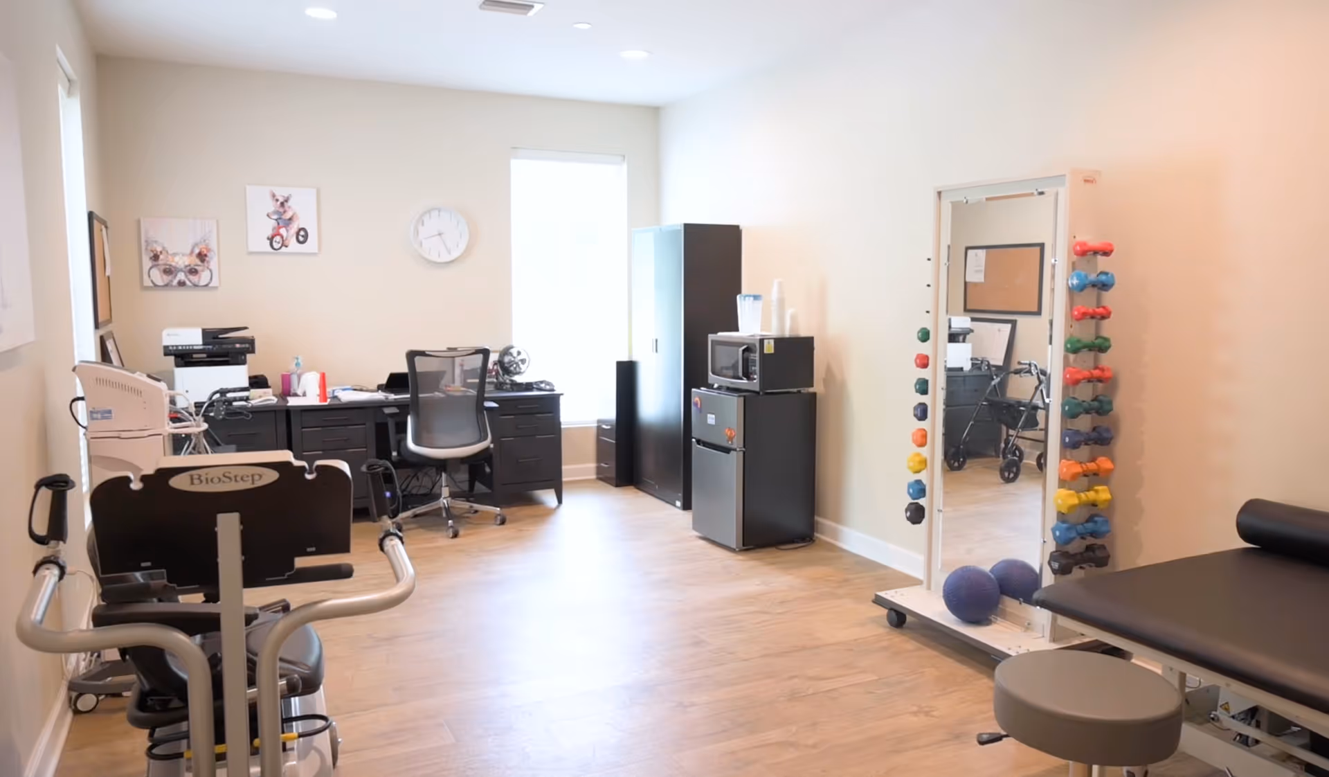 A bright room with light wood flooring featuring exercise equipment including a BioStep machine, a rack of colorful dumbbells next to a full-length mirror, a black padded examination or therapy table with a stool, and a desk with office supplies and a chair near a window. There is also a small refrigerator and microwave on a cabinet against the wall.
