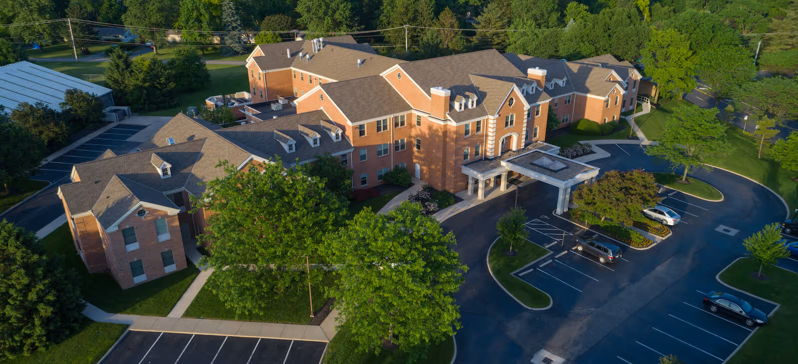 Aerial view of the Harmony Trace senior living facility showing a large brick building with a covered entrance, parking lot, and surrounding trees.