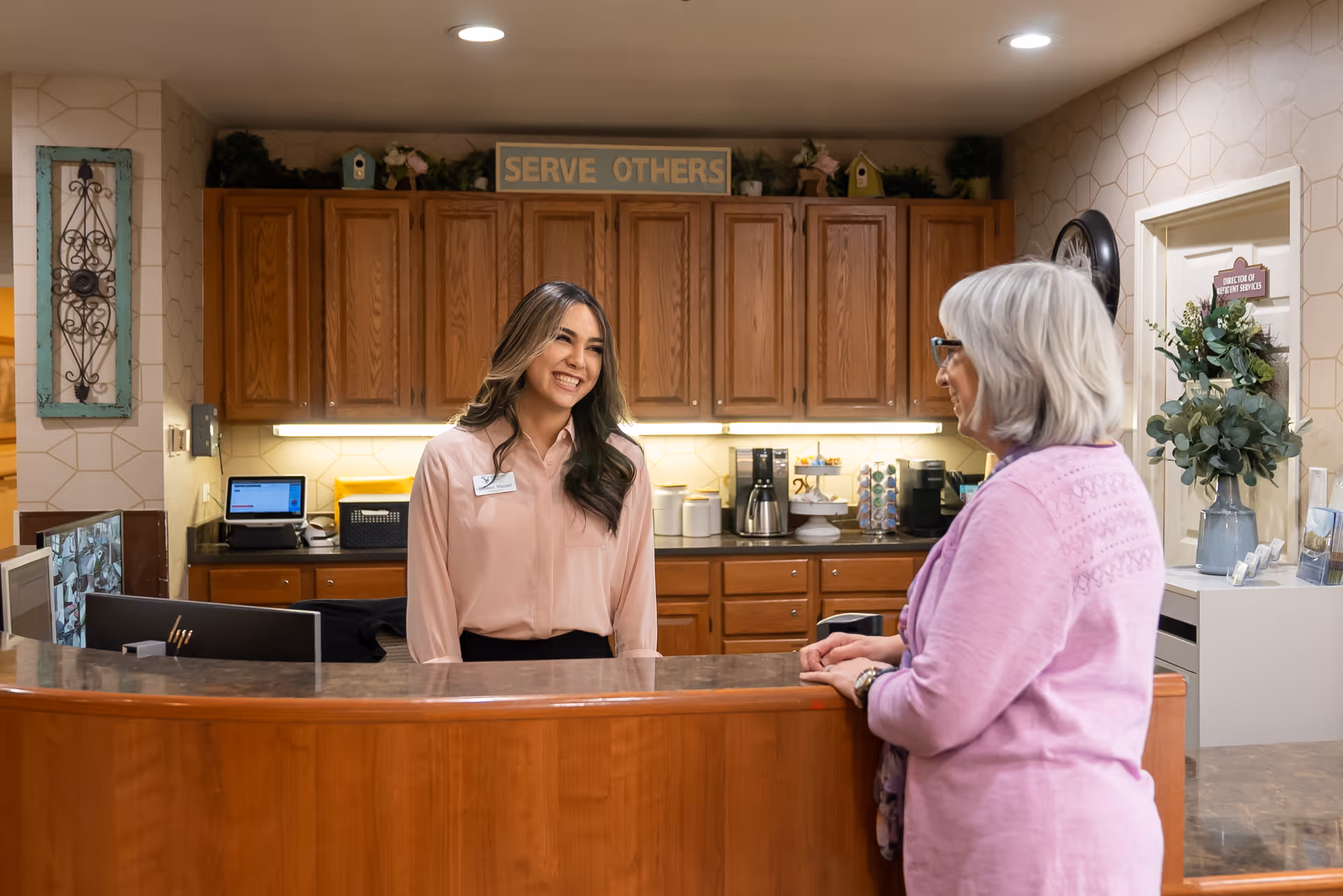A smiling staff member stands behind a reception desk talking with a resident in a senior living facility lobby.
