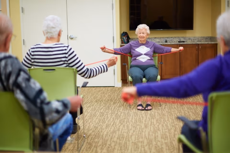 A group of elderly individuals seated in a room participating in a seated exercise session using red resistance bands. One elderly woman in a purple sweater is leading the exercise, stretching the band with her arms extended. The room has beige walls, a carpeted floor, green chairs, and a flat-screen TV mounted on the wall.