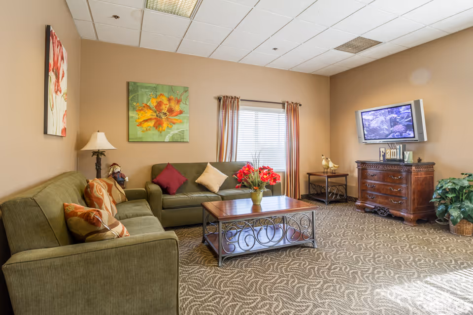 A cozy living room with two green sofas adorned with colorful throw pillows, a wooden coffee table with a decorative vase of red flowers, a window with striped curtains, a wooden dresser with a flat-screen TV mounted above it, and wall art featuring floral designs. The room has beige walls and patterned carpet flooring.