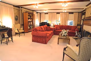 A cozy living room with a large red sectional sofa, a wooden coffee table with a floral centerpiece, several upholstered chairs, a wooden bookshelf, and a fireplace. The room has beige carpeting, cream-colored curtains, and warm lighting.