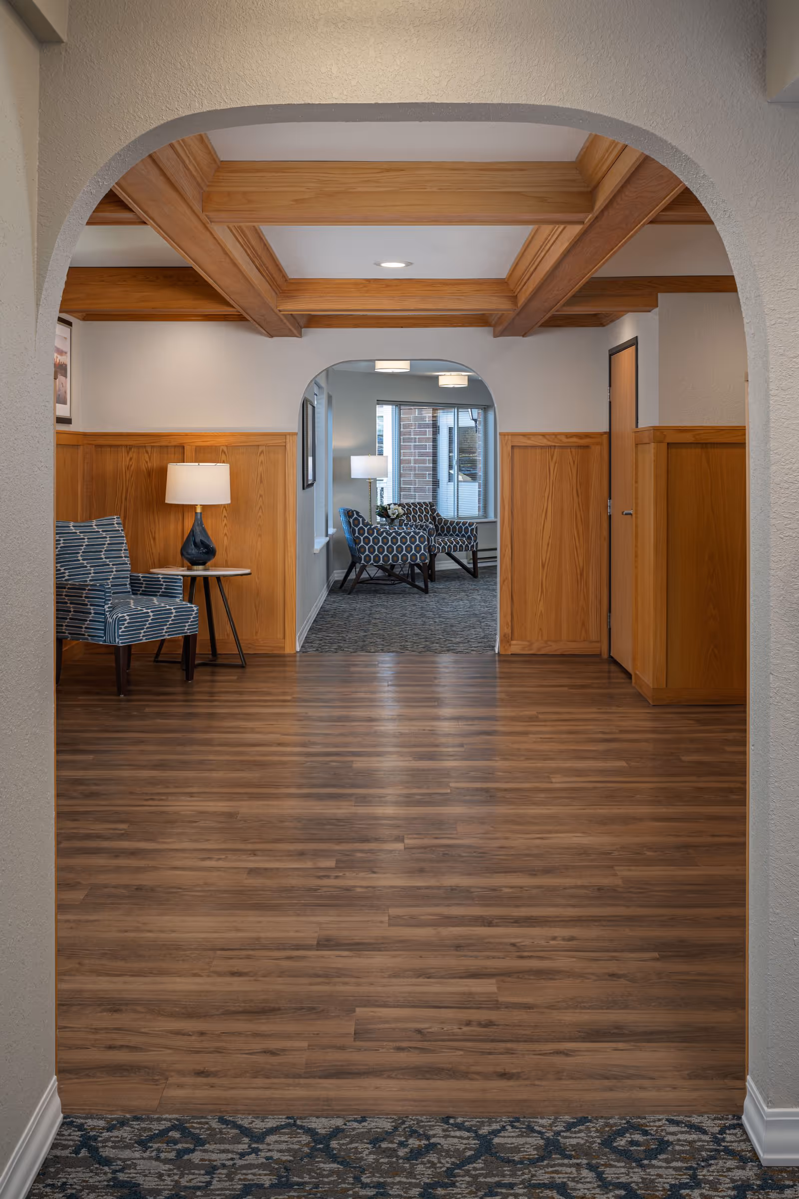 View through an arched doorway into a senior living facility interior featuring wood-paneled walls and a coffered ceiling. The foreground has a wood floor leading to a carpeted sitting area with patterned armchairs and a side table with a lamp. Large windows in the background let in natural light.