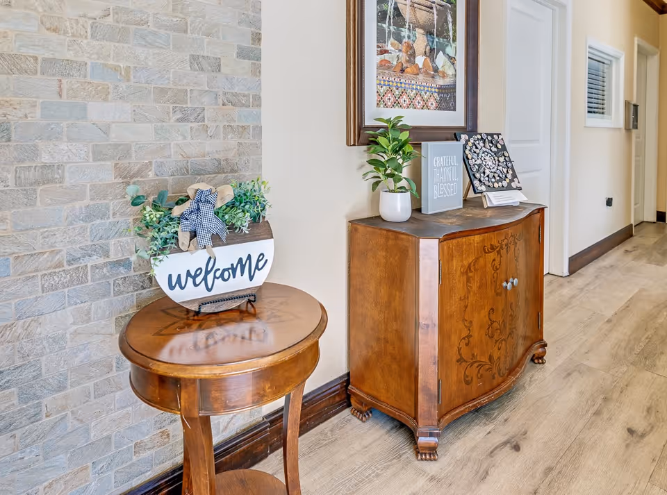 A hallway corner in a senior living facility featuring a small round wooden table with a decorative 'welcome' sign and greenery, next to a wooden cabinet with intricate designs. On top of the cabinet are a potted plant, a framed picture, and a sign that reads 'Grateful Thankful Blessed'. The wall behind has a textured stone finish and a framed artwork above the cabinet. The floor is light wood, and there are doors and a window along the hallway.