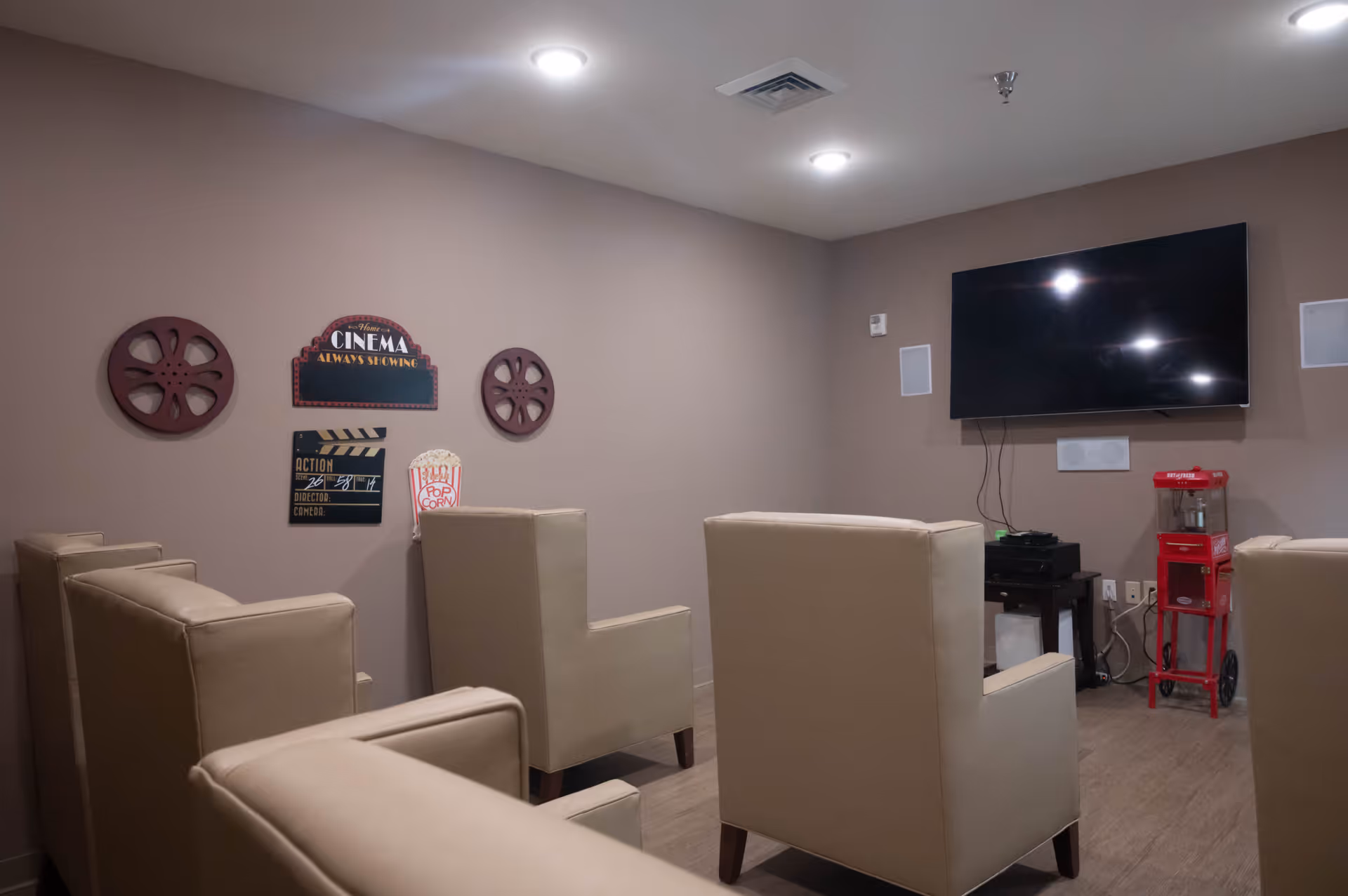 A small movie theater room with beige armchairs arranged in rows facing a wall-mounted flat-screen TV. The walls are decorated with cinema-themed signs and film reels. A red popcorn machine is positioned near the TV.