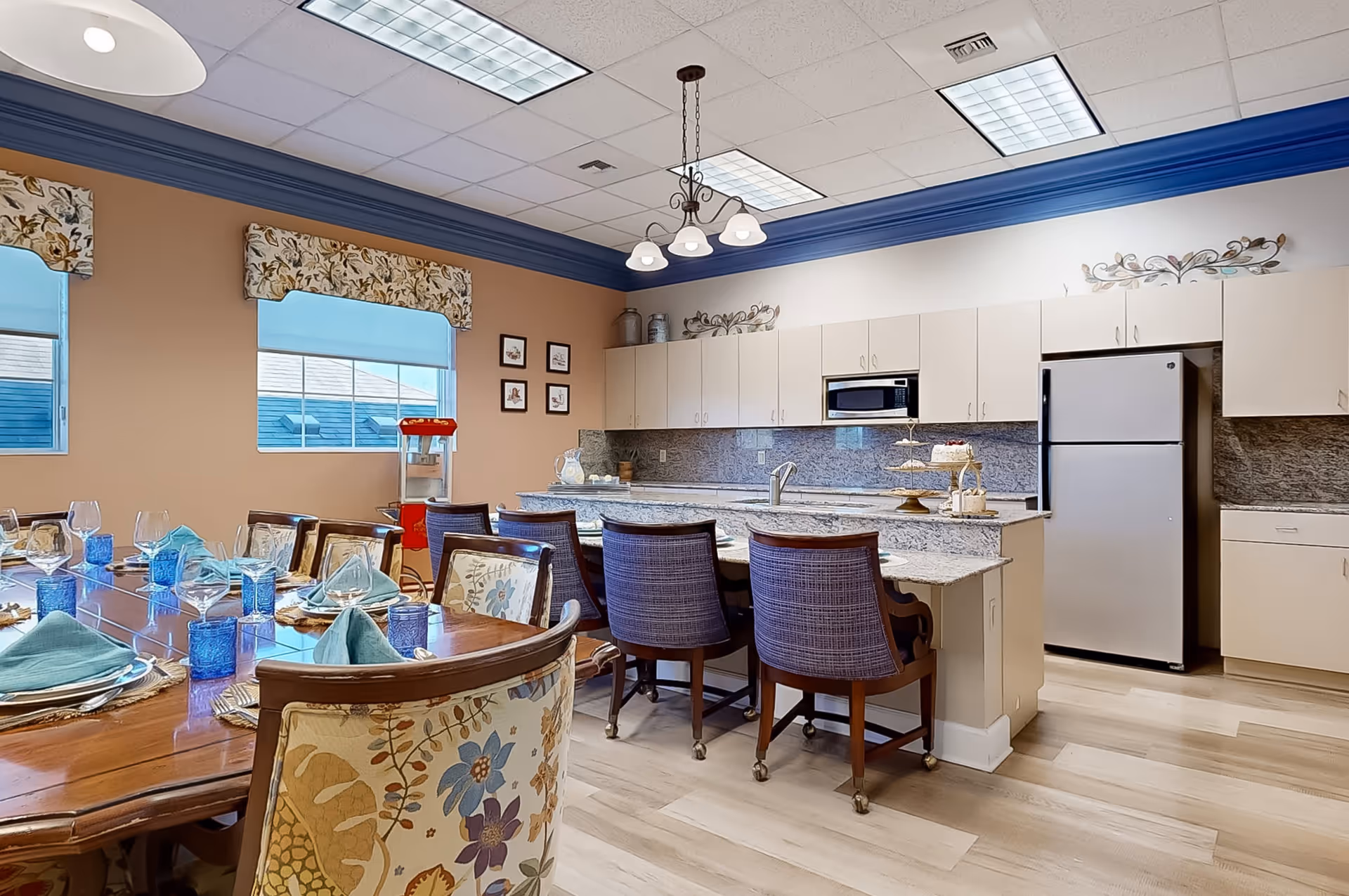 A well-lit kitchen and dining area with a large wooden dining table set with blue glasses, plates, and napkins. The kitchen features white cabinets, a granite countertop island with four chairs, a refrigerator, microwave, and a popcorn machine near the window. The walls are painted beige with blue crown molding and floral valances on the windows.