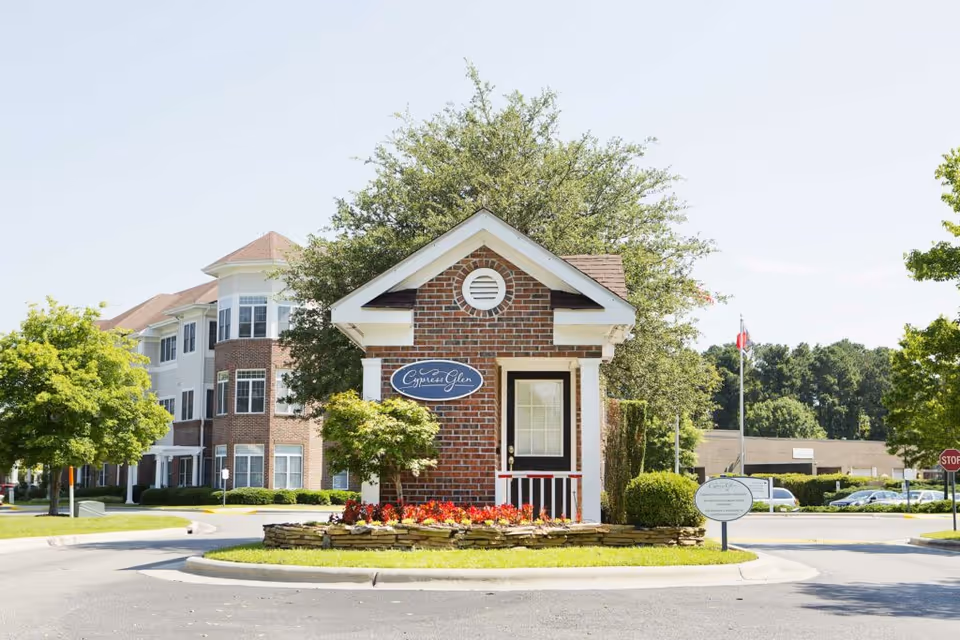 Small brick guardhouse with a sign reading 'Cypress Glen' in front of a large multi-story residential building surrounded by trees and landscaping under a clear sky.