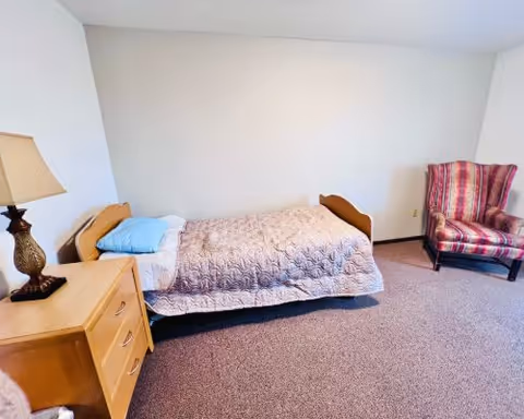 Small furnished bedroom with a single bed, wooden nightstand and lamp, and a striped upholstered armchair against a plain light-colored wall.