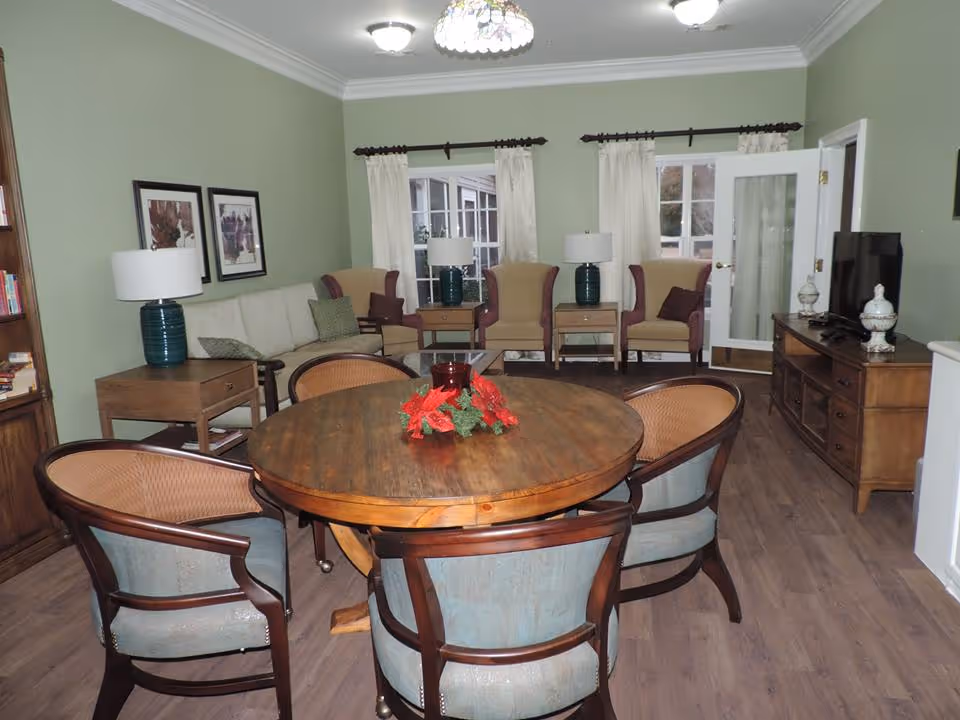 A cozy senior living common area with a round wooden table surrounded by six cushioned chairs. The room has light green walls, wooden flooring, and white curtains on windows. There are three armchairs and a sofa along the far wall, two table lamps with blue bases on side tables, framed artwork on the wall, and a wooden TV stand with a flat-screen TV and decorative items.