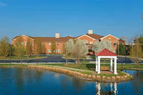 Brick senior living facility by a pond with a gazebo and trees.