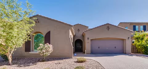 Front exterior view of a single-story stucco house with a two-car garage, arched windows, and a small landscaped area with trees and shrubs under a clear blue sky.