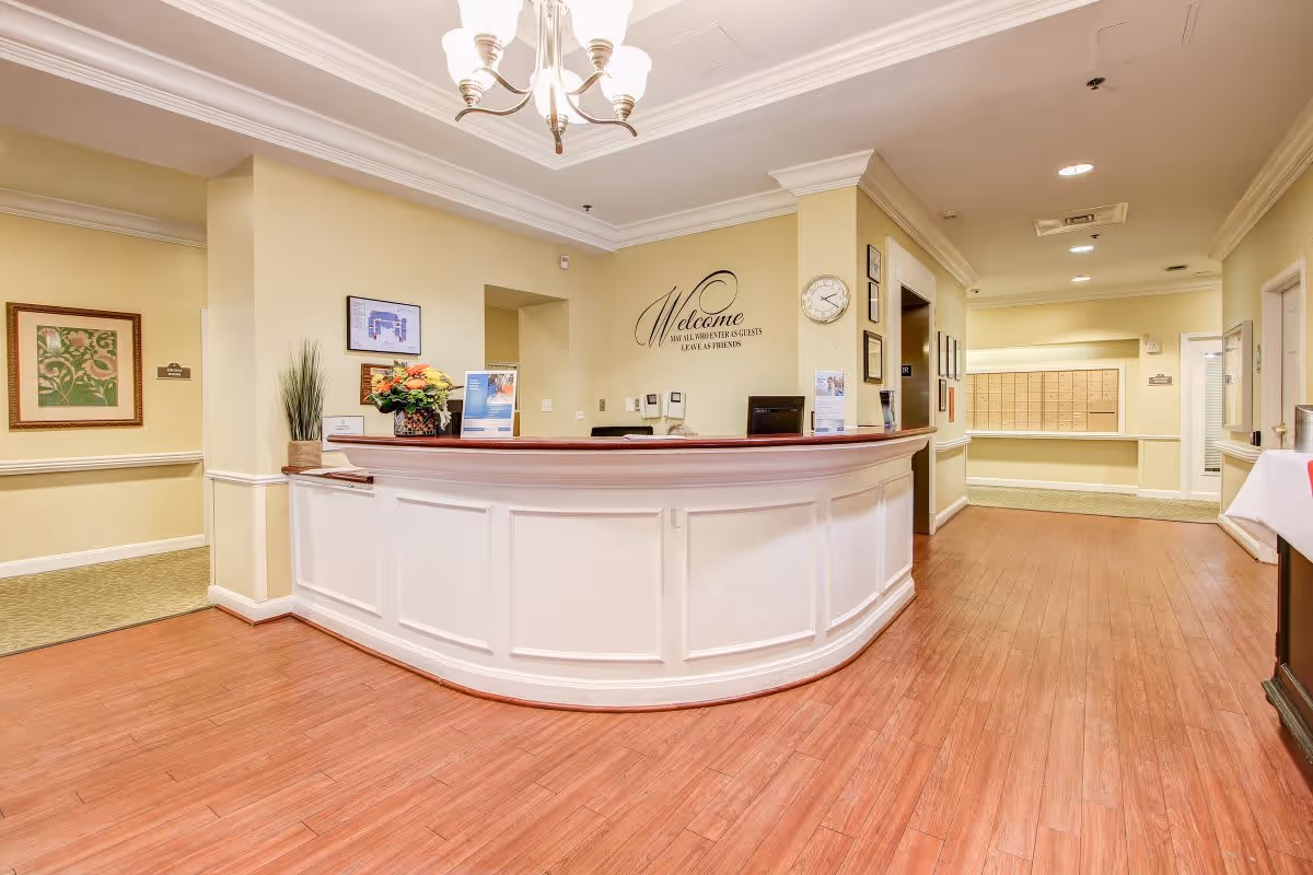 Reception area of a senior living facility with a white curved front desk, a chandelier overhead, a clock on the wall, framed artwork, and a welcoming sign that reads 'Welcome. You are all visitors & guests. Leave as friends.' There are mailboxes visible in the background along a hallway with wooden flooring and cream-colored walls.