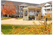 Outdoor courtyard area of a senior living facility with a wooden pergola, tables and chairs, surrounded by a three-story building with balconies. There are autumn-colored trees and orange flowers in the foreground.