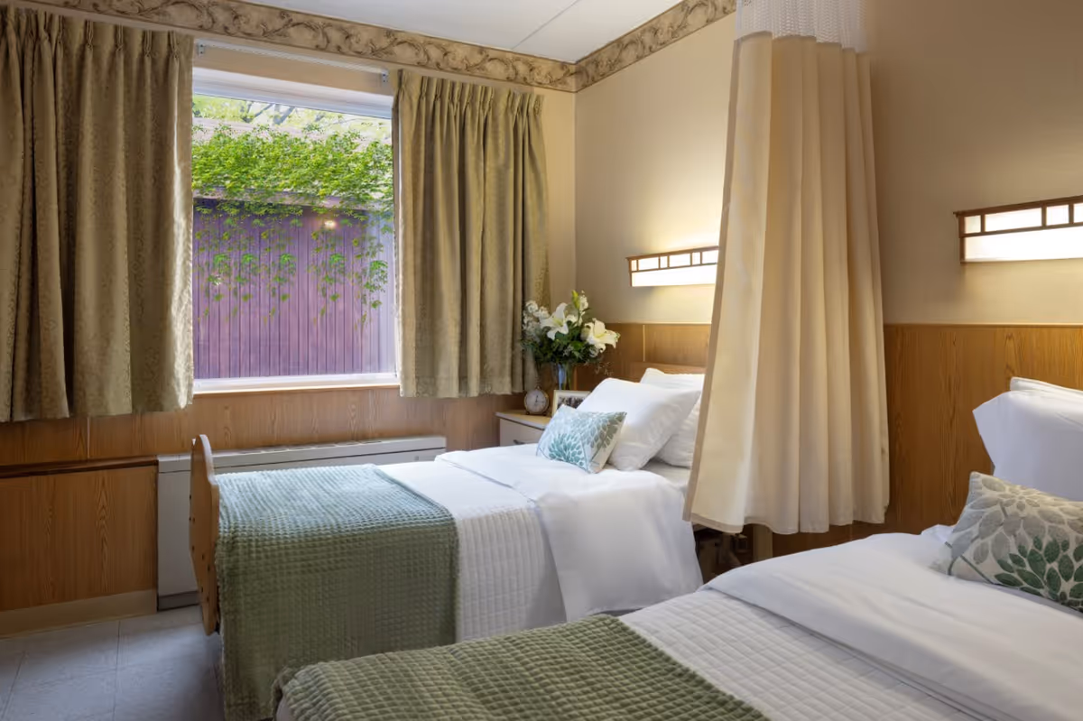 Two twin beds in a bright senior-living bedroom with green quilts, a window with curtains, lamps, and a privacy curtain between the beds.