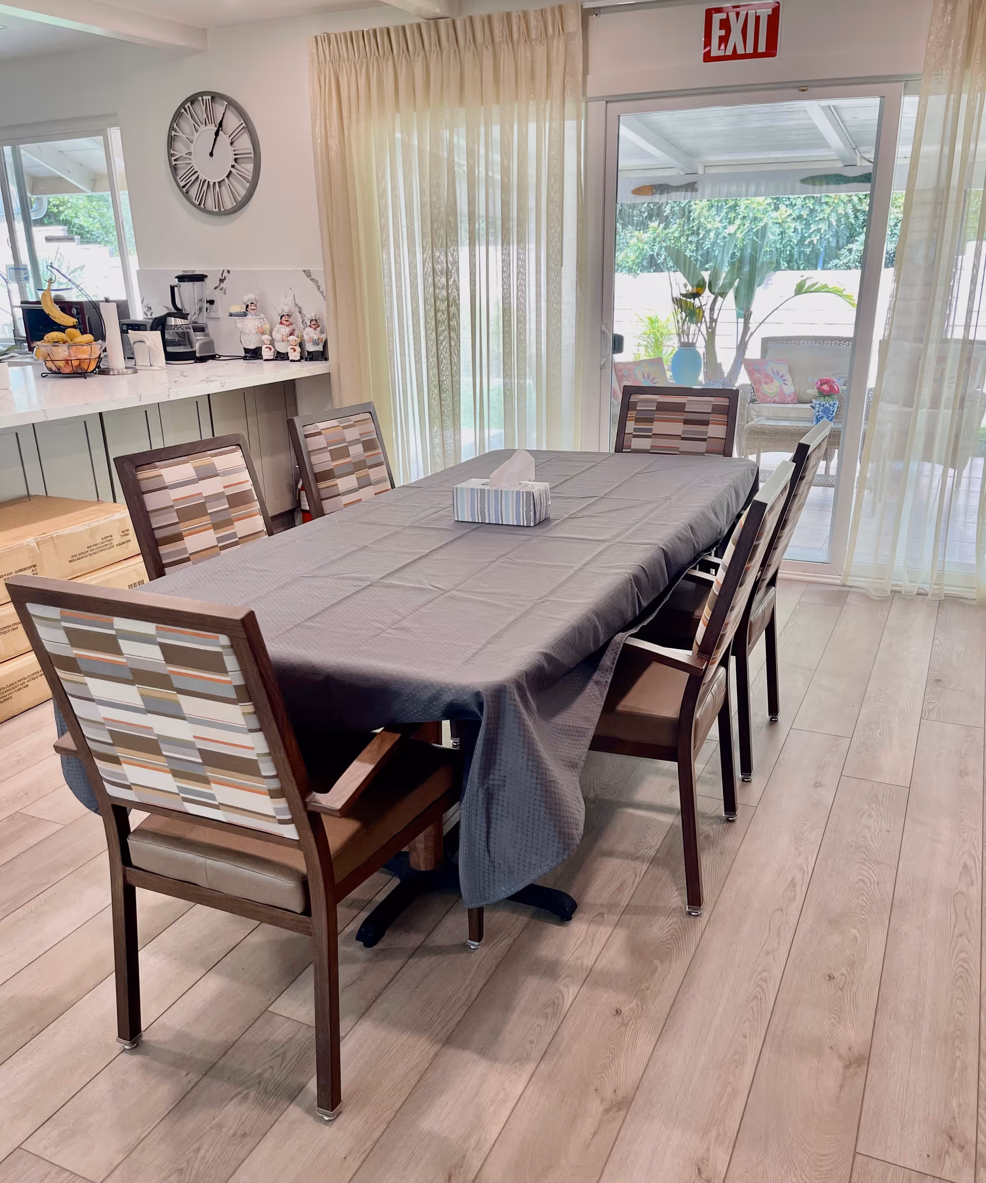 A dining area with a rectangular table covered by a gray tablecloth and six chairs with patterned backs. Behind the table, there is a sliding glass door with sheer curtains leading to an outdoor patio. To the left, a kitchen counter with a blender, coffee maker, and decorative items is visible. A large wall clock is mounted above the counter, and an exit sign is above the sliding door.