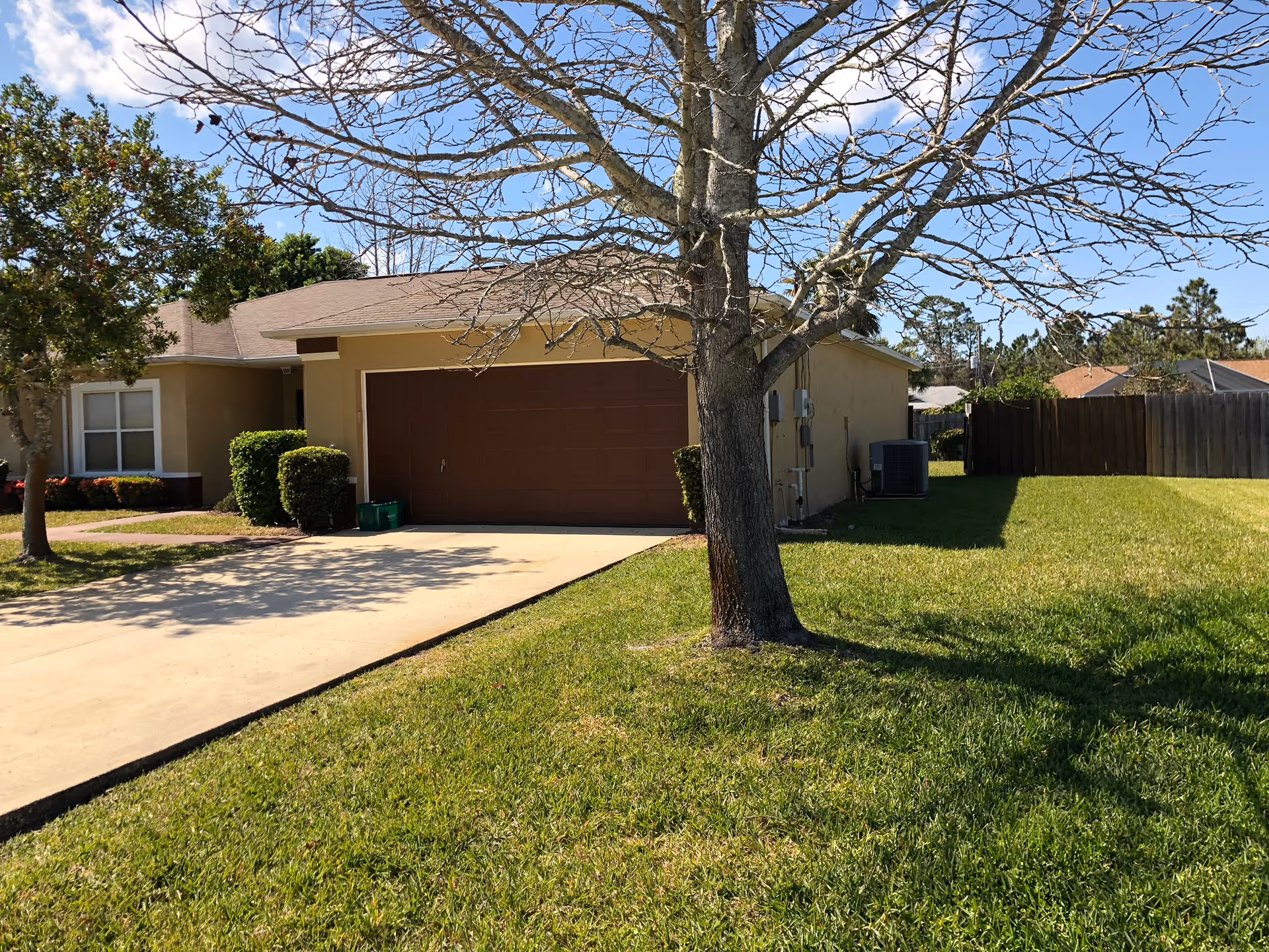 Single-story house with a brown garage door, beige exterior walls, and a sloped roof. In front of the house is a concrete driveway and a grassy lawn with a leafless tree casting shadows. There are some bushes near the house and a wooden fence in the background under a clear blue sky.