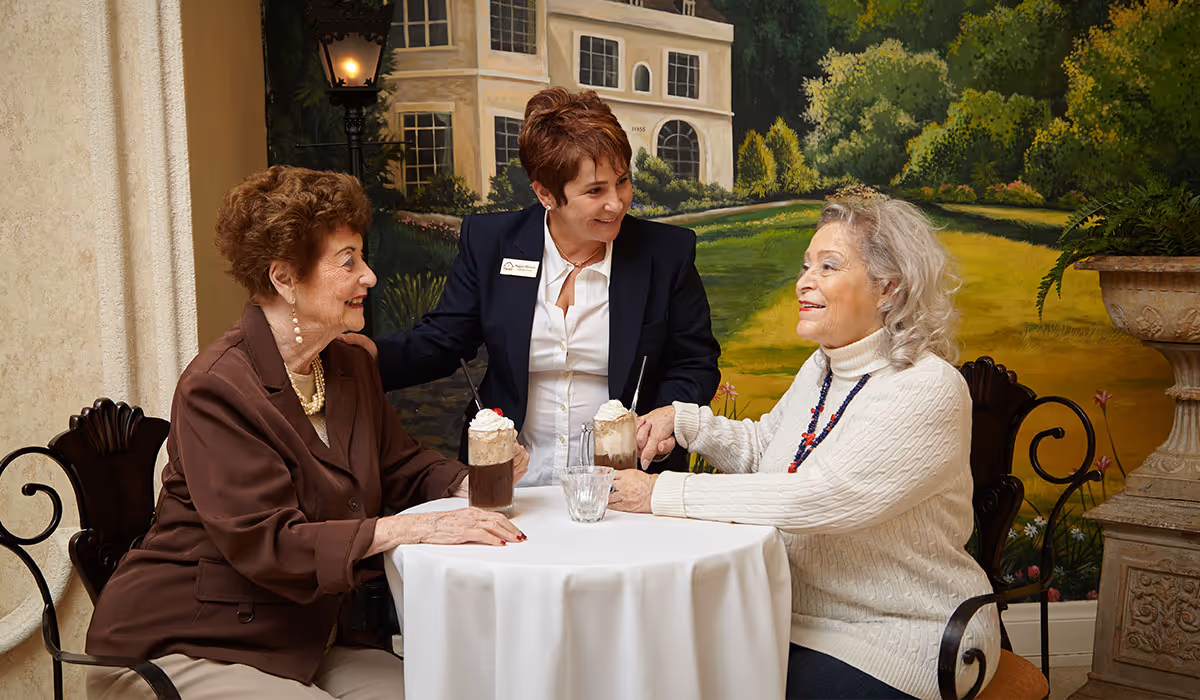 Two elderly women sitting at a round table with a white tablecloth, each with a glass of iced coffee topped with whipped cream, while a staff member in a dark blazer and white shirt stands between them, smiling and holding their hands. The background features a mural of a large house and greenery.
