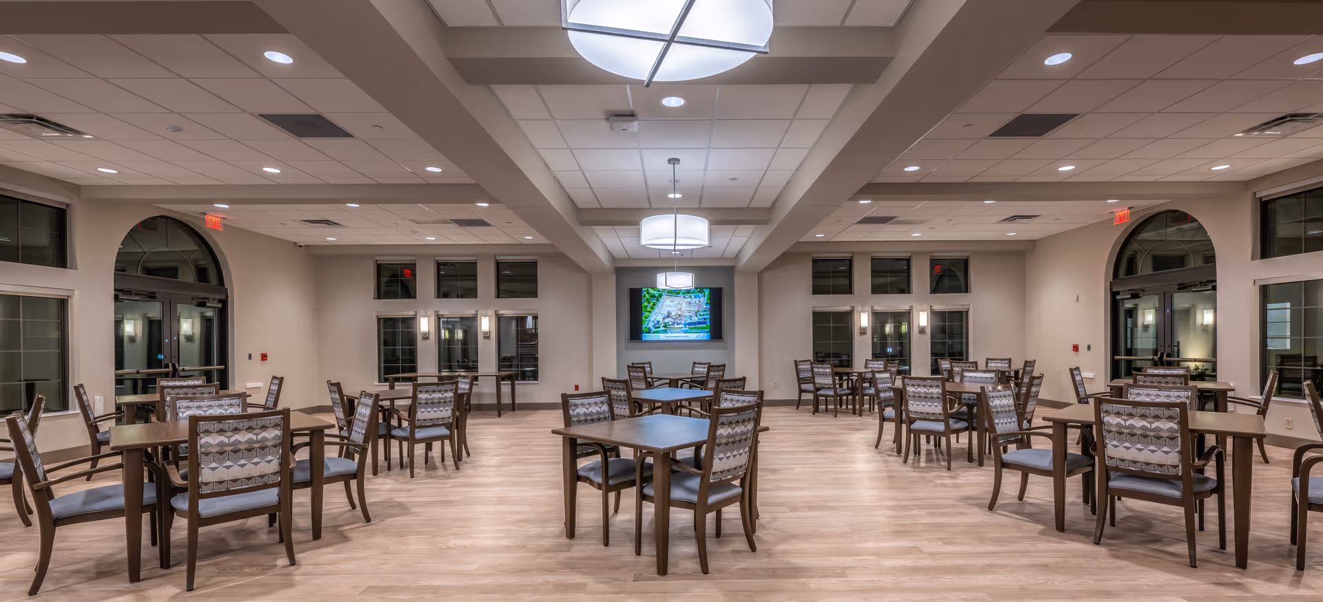 A spacious dining room with multiple square wooden tables and cushioned chairs arranged neatly. The room has large windows, light-colored walls, and a light wood floor. Ceiling lights provide bright illumination, and a flat-screen TV is mounted on the far wall.