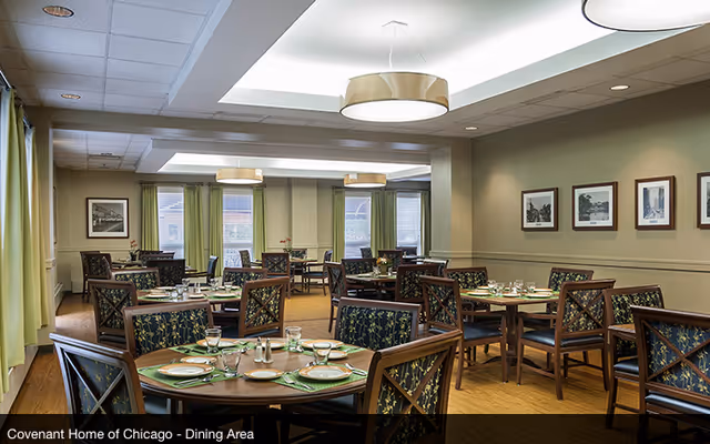 A dining area with multiple round and square wooden tables set with plates, glasses, and silverware. The chairs have dark wooden frames with patterned upholstery. The room has light green walls, large windows with green curtains, and framed black and white photographs on the wall. Ceiling lights provide soft illumination.