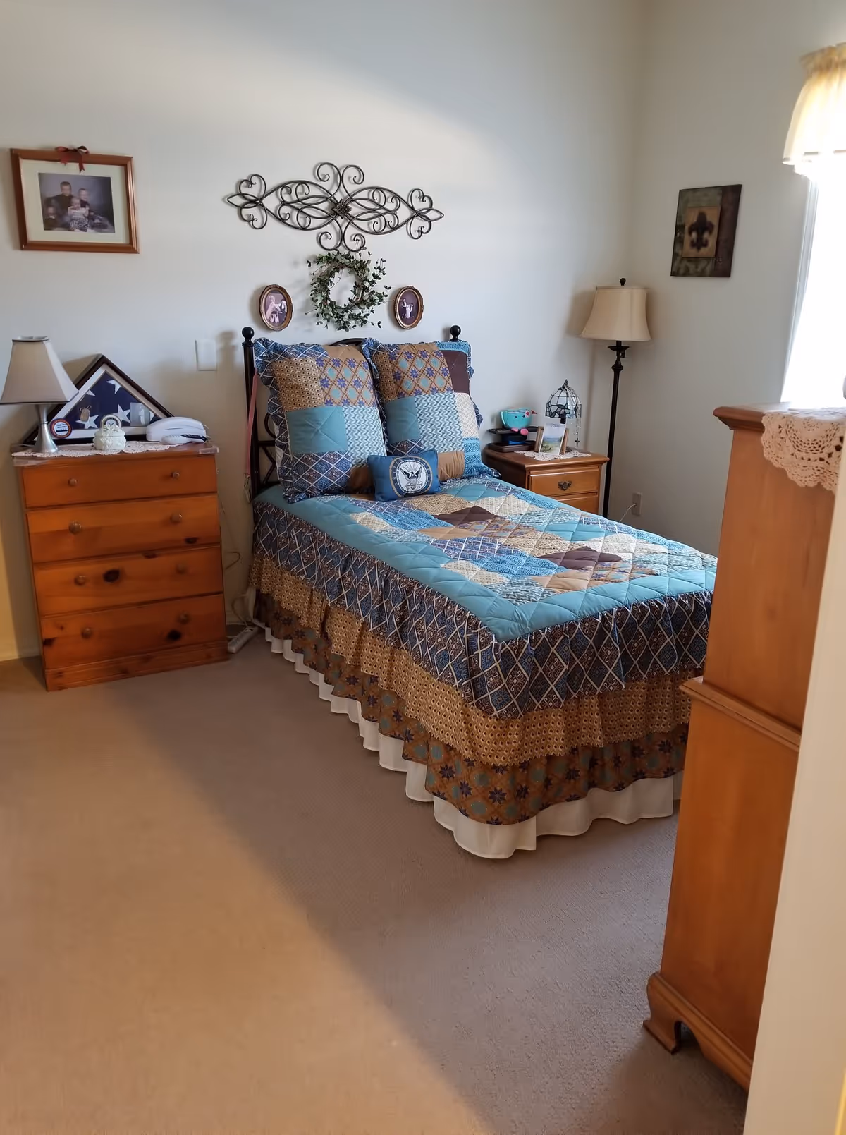 A neatly arranged bedroom with a twin bed covered in a blue, brown, and beige patchwork quilt. The bed has matching pillows and a small decorative pillow with an emblem. On either side of the bed are wooden nightstands with lamps and various small items. Above the bed is a decorative metal wall art piece and two small framed pictures. The room has beige carpet and light-colored walls with a window covered by a sheer curtain letting in natural light.