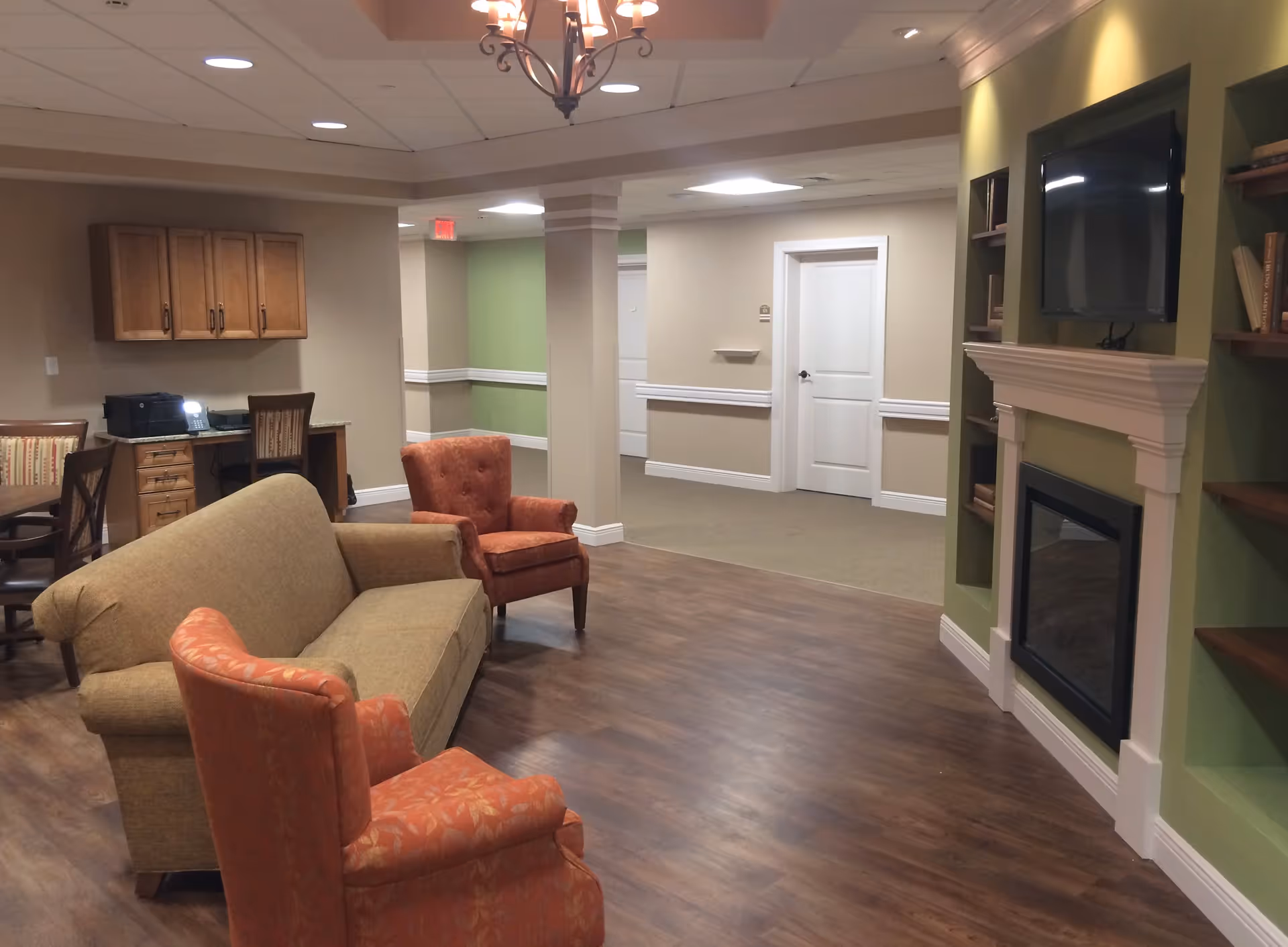 A cozy living room area in a senior living facility with a beige sofa, two orange armchairs, a wall-mounted TV above a white fireplace, built-in shelves with books, a wooden floor, and a small desk with a chair and printer in the background.