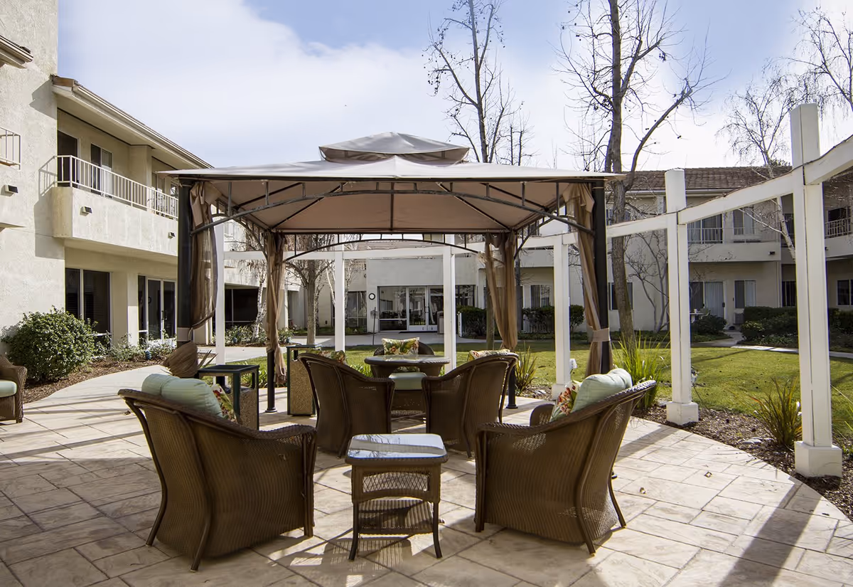 Outdoor seating area at Sunrise At Sterling Canyon with a canopy-covered patio, wicker chairs with cushions, a small table, and surrounding greenery and buildings under a partly cloudy sky.