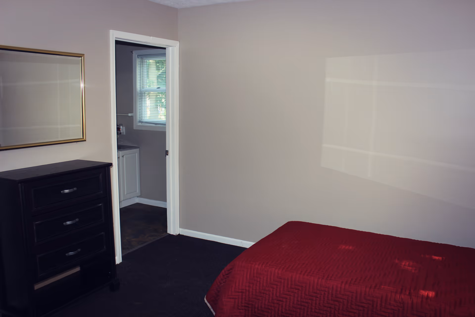 Simple bedroom with a red bedspread, a dark dresser and an open door showing a small bathroom sink and window.