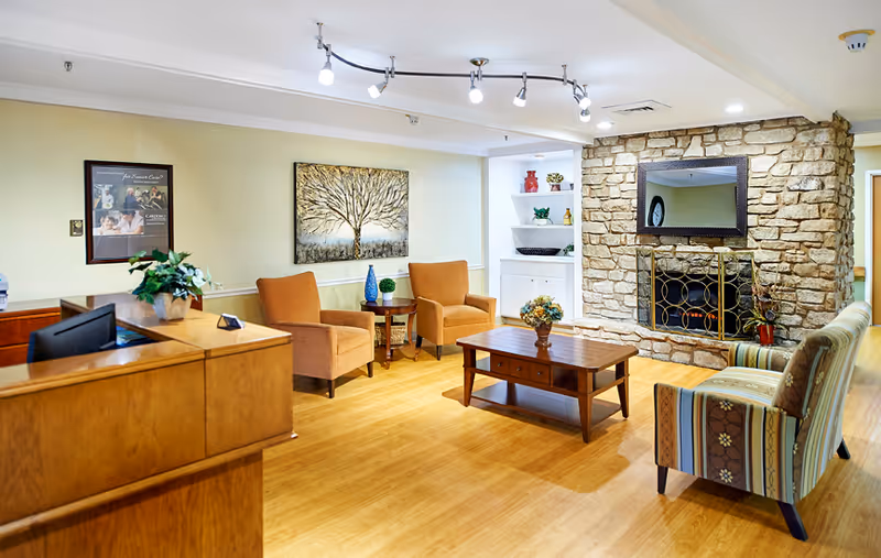 Bright communal living area with a reception desk, armchairs, a coffee table, shelving, and a stone fireplace.