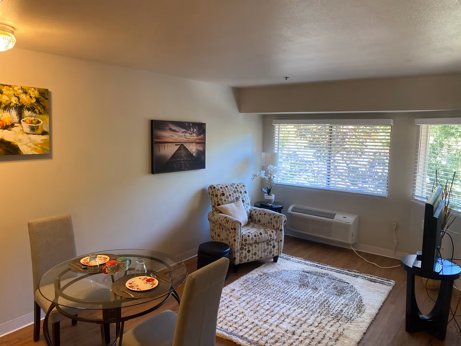 A cozy living area with a glass-top round dining table set for two, two beige upholstered chairs, a patterned armchair with a pillow, a side table with a lamp and a potted orchid, a large window with blinds, a wall-mounted air conditioning unit, a flat-screen TV on a stand, and two paintings on the walls. The floor has a light-colored patterned rug and wood flooring.