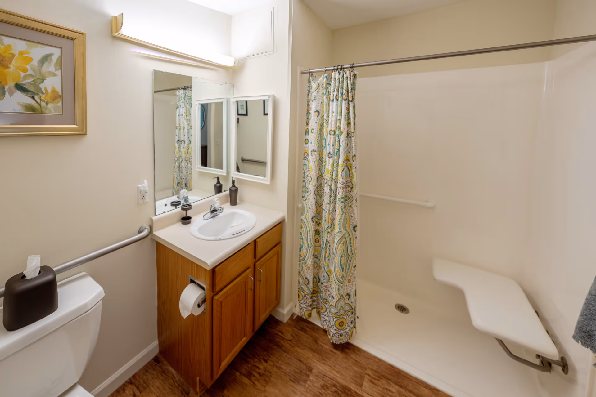 A bathroom with a toilet, a wooden vanity with a sink, a mirror above the sink, a floral shower curtain, and a walk-in shower with a fold-down seat. There is a grab bar next to the toilet and another inside the shower. A framed floral picture hangs on the wall above the toilet.