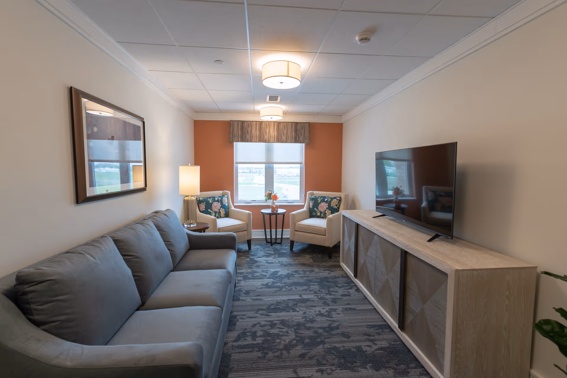 A cozy living room with a gray sofa on the left, two beige armchairs with floral cushions near a window with an orange accent wall, a small round table with a flower vase between the chairs, a wooden TV stand with a flat-screen television on the right, and a framed picture on the left wall.