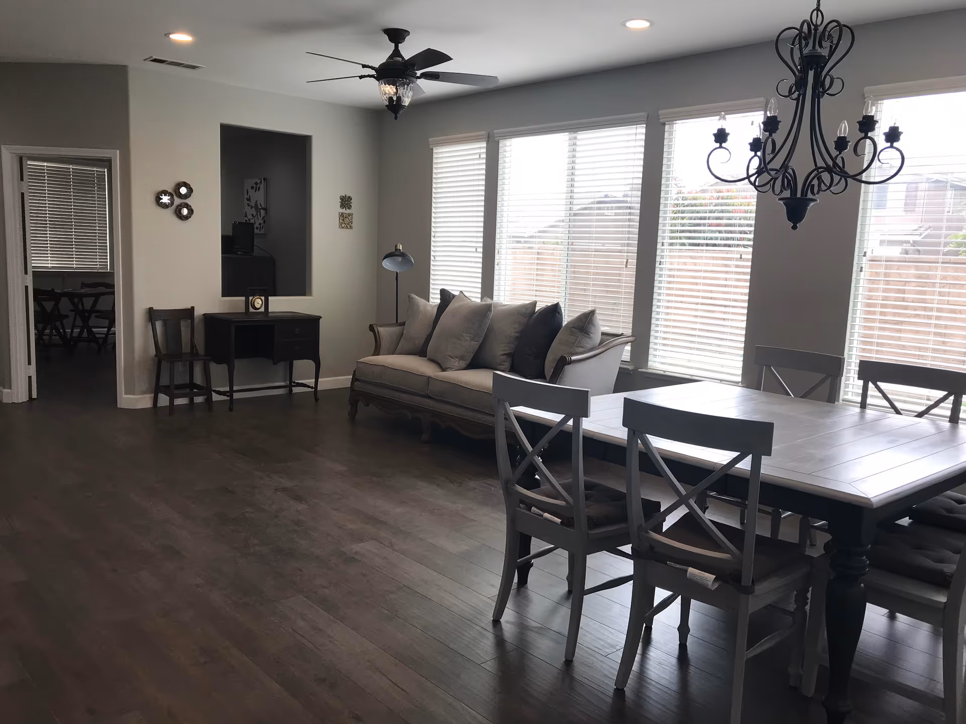 A bright interior room featuring a dining area with a rectangular table and six chairs, a beige sofa with multiple cushions, a ceiling fan with light, a chandelier, and large windows with blinds letting in natural light. There is also a small desk with a chair and decorative wall hangings.