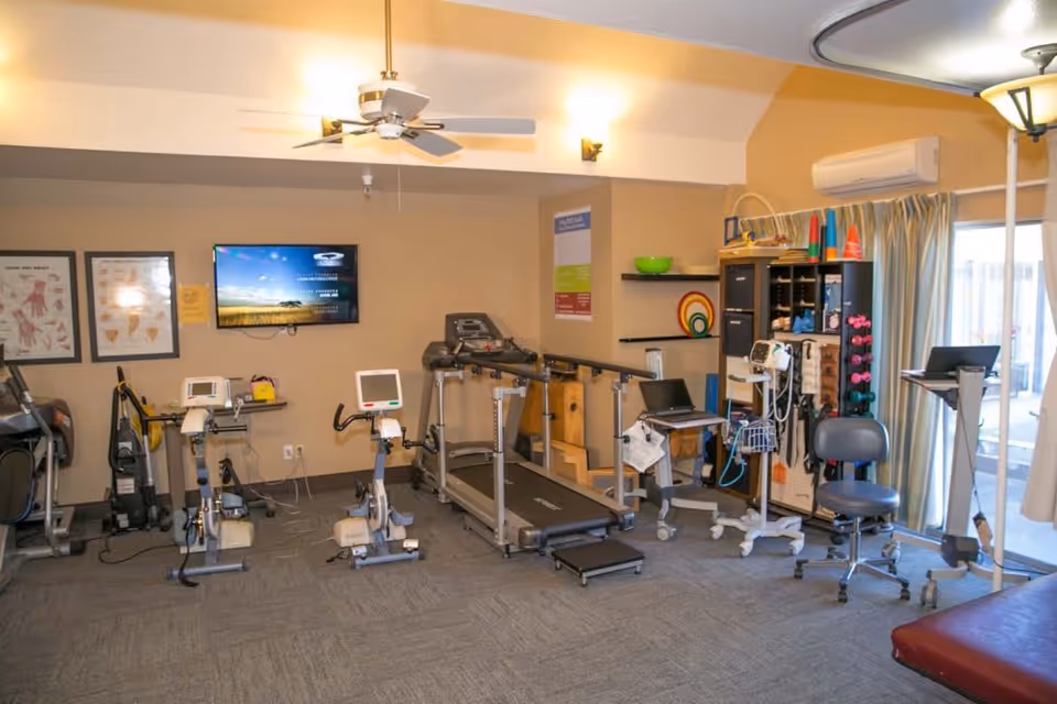 A rehabilitation or physical therapy room with exercise equipment including stationary bikes, a treadmill with handrails, medical monitoring devices, and various therapy tools. The room has beige walls, a ceiling fan, a wall-mounted TV, and a window with curtains allowing natural light.