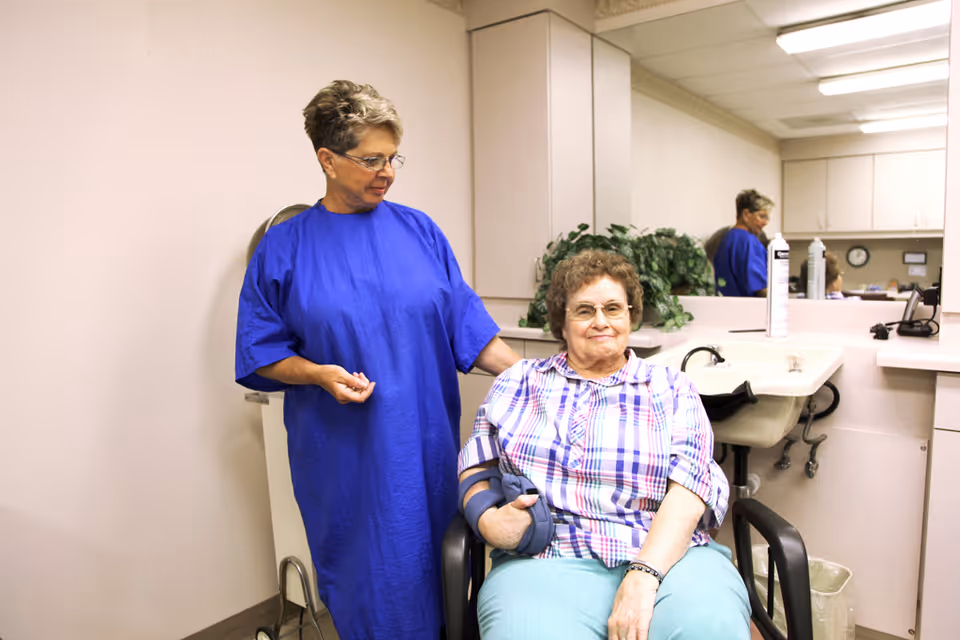 An elderly woman sitting in a wheelchair with her arm in a sling, accompanied by a caregiver wearing a blue gown standing beside her in a room with a sink, mirror, and cabinets.
