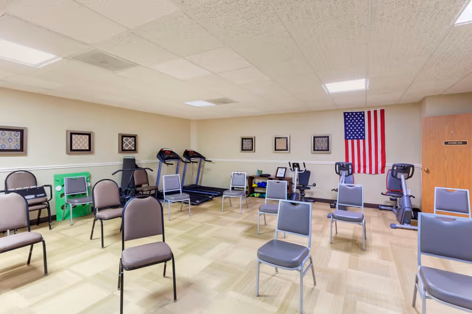 A spacious exercise room with several chairs arranged in rows, two treadmills, two stationary bikes, and exercise equipment on shelves. An American flag hangs on the wall next to a door labeled 'EXERCISE'. The room has beige walls, framed artwork, and a tiled ceiling with fluorescent lights.