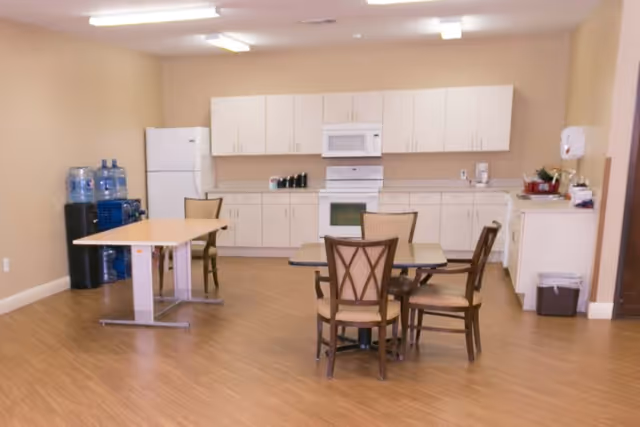 A kitchen and dining area with light wood flooring, beige walls, and white cabinetry. The kitchen features a white refrigerator, microwave, and stove. There are two tables with chairs around them, and a water dispenser with large water bottles is visible in the corner.